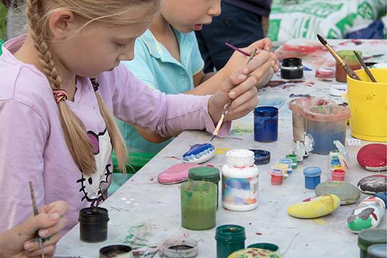 Young girl with blonde pigtails paints a rock at a table of peers.